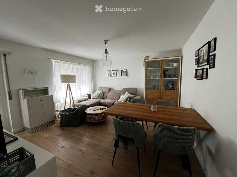 A modern living room with a sectional sofa, coffee table, dining table, chairs, and a cabinet. The room features wooden flooring, large windows with curtains, and several picture frames on the walls.