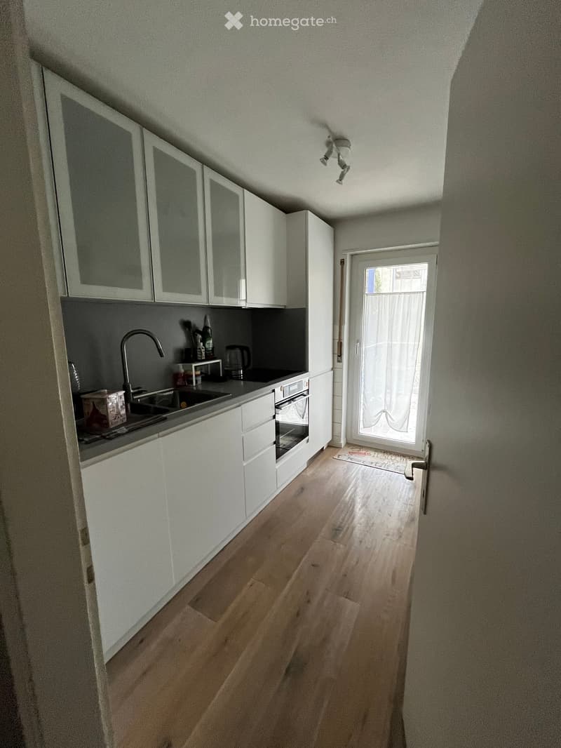 Modern kitchen with white cabinets, wooden flooring, and a sink with a faucet. A small window with a curtain is visible, and the space is well-lit with ceiling lights.