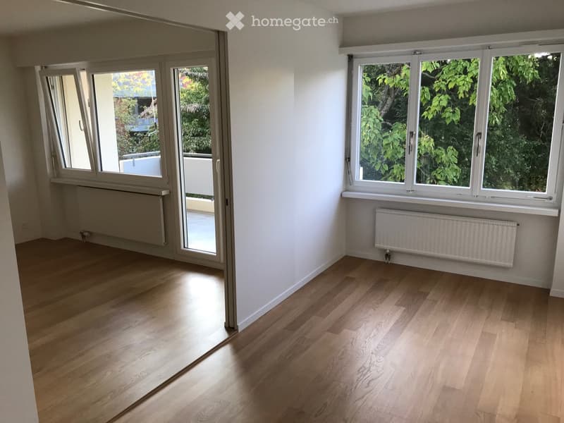 Empty room with light wooden flooring, white walls, large windows, and a sliding glass door leading to a balcony.