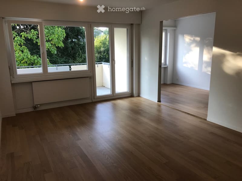 Empty room with wooden flooring, large windows, and a sliding glass door leading to a balcony area.