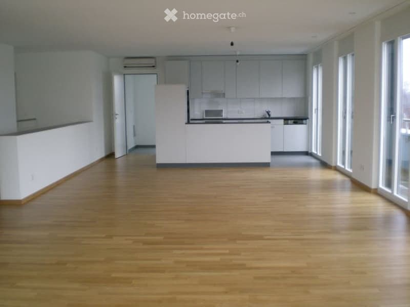 Empty room with wooden flooring, white walls, and a white kitchen area in the background. No furniture or equipment is present.