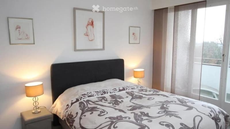 Modern bedroom with a large bed, nightstands, table lamps, and framed artwork on the walls. Large windows with sheer curtains allow natural light to enter.