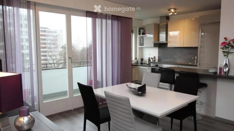 Modern, open-plan living and dining area with a white dining table, chairs, and a kitchen area featuring cabinets, an oven, and a dishwasher. Large windows with sheer curtains offer a view of the balcony.