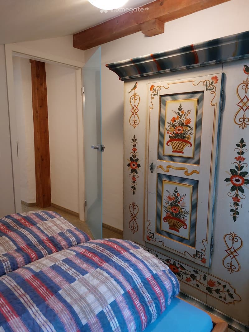 A room with a wooden cabinet with floral design and two beds covered with multicolored blankets. Behind the bed, there is a wooden pillar next to an open glass door. On top, there is a ceiling lamp