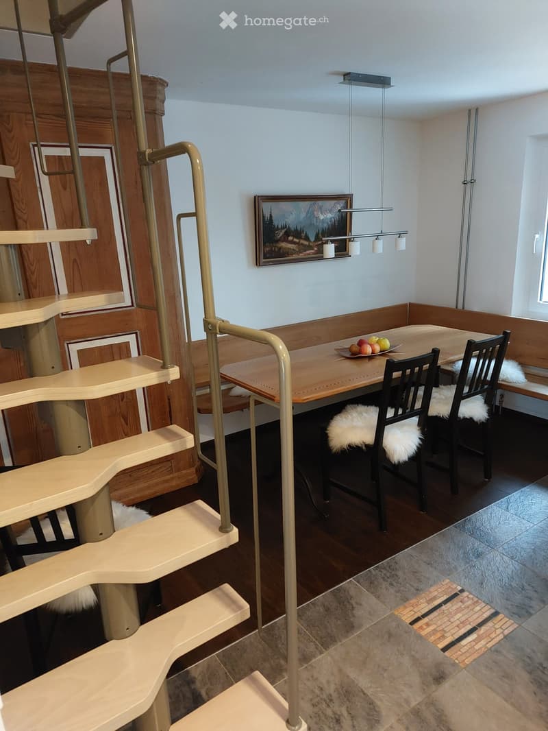 Dining area with wooden table, chairs, L-shaped bench, hanging lamps, wall mounted picture frame, staircase