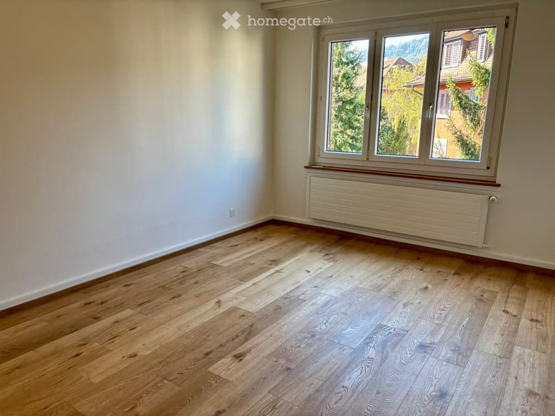 Empty room with light wooden flooring, white walls, and a large window with a radiator below it.