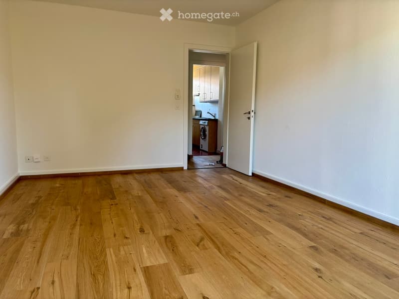 Empty room with light wooden flooring and white walls, featuring an open doorway leading to a small kitchen area with cabinets and a washing machine.