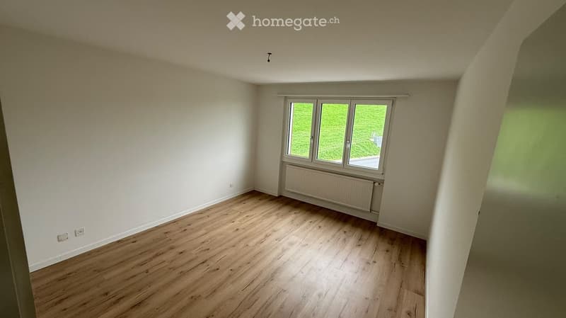 Empty room with wooden flooring, white walls, and a window with a view of greenery outside.
