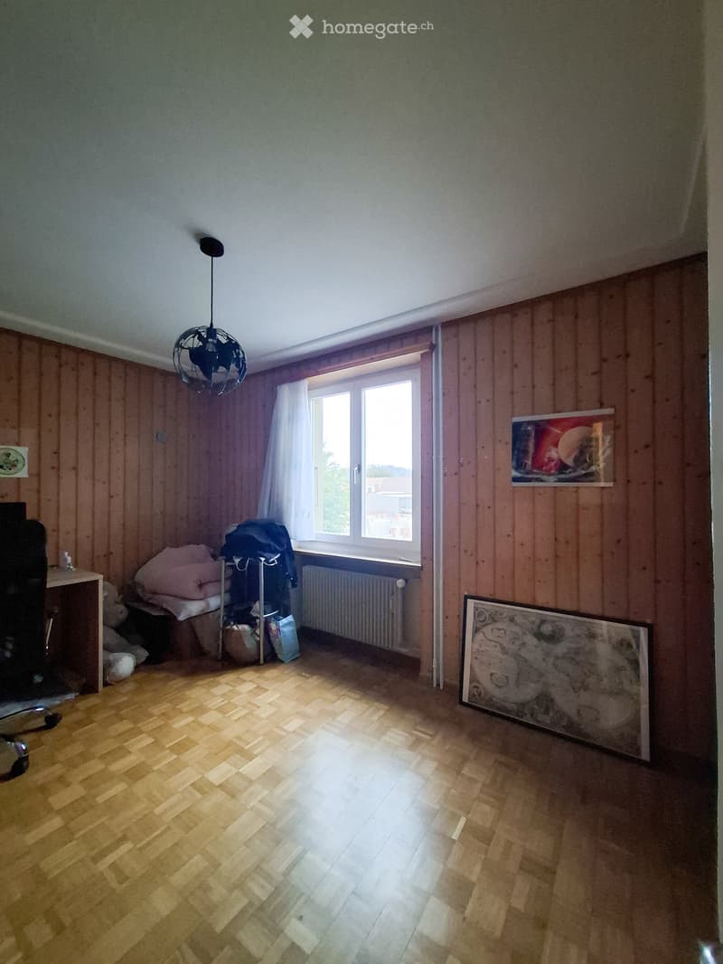 A furnished room with wooden panel walls, a large window with white curtains, a wooden floor, a chair, a small table, and picture frames on the wall.