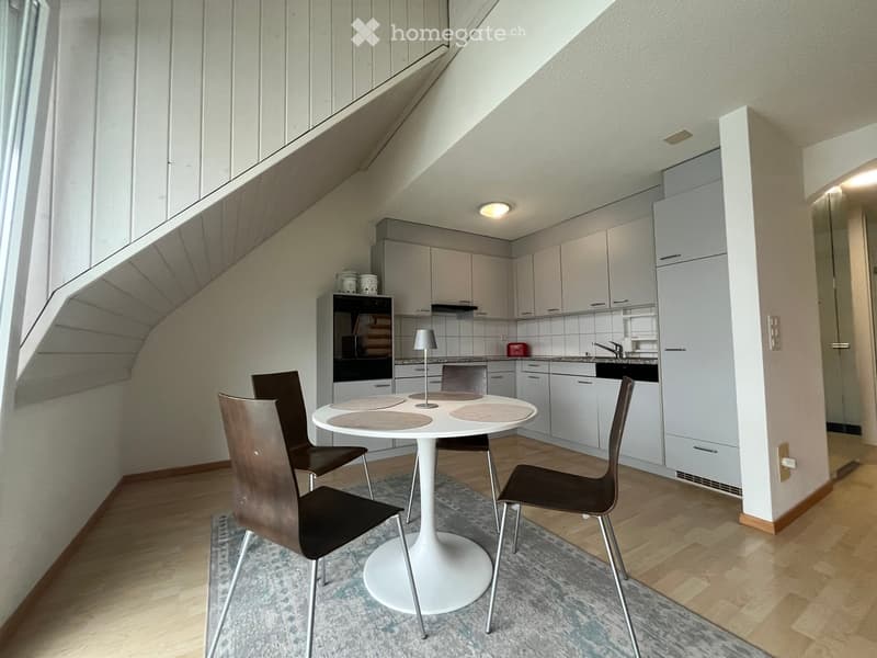 A modern, compact kitchen and dining area with white cabinets, a white round table, four chairs, and an oven. The space features wooden flooring and a skylight providing natural light.