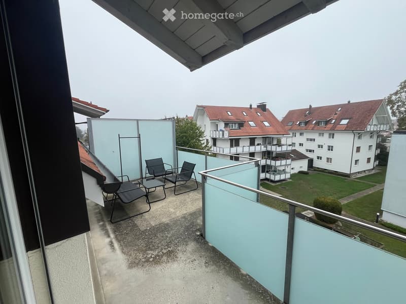 Balcony with modern glass railing, featuring two black chairs and a small table, overlooking a residential area with houses and greenery.