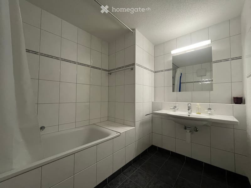 Modern bathroom with a bathtub, sink, and mirror. The walls are tiled in light gray with dark gray tiles on the floor.