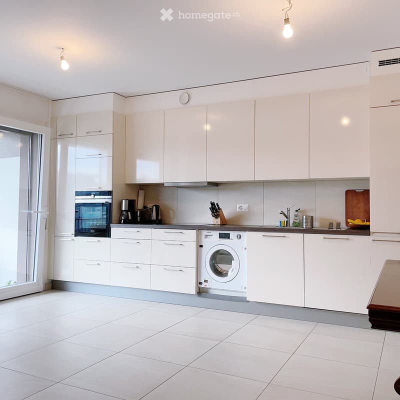 Modern kitchen with white cabinets, tiled floor, and stainless steel appliances including an oven, washing machine, and coffee maker.