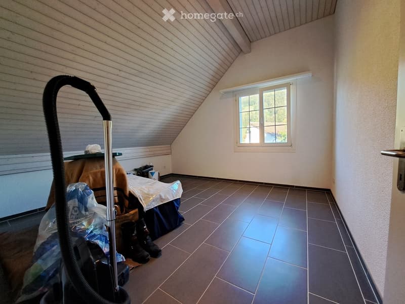 Empty room with tiled floor, slanted wooden ceiling, and a window with white frame. A vacuum cleaner is partially visible in the foreground, and some items are stacked near the window.