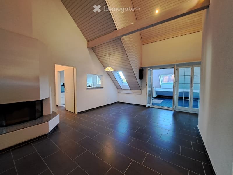An empty room with dark tiled flooring, white walls, and a slanted wooden ceiling. Features include a fireplace on the left, a doorway leading to another room, and large glass doors opening to a balcony or outdoor area.