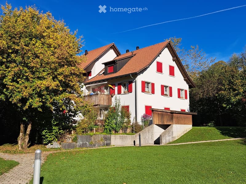 Two-story house with red shutters, red-tiled roof, and a raised ground floor. The house features a wooden balcony and is surrounded by a well-maintained lawn and trees.