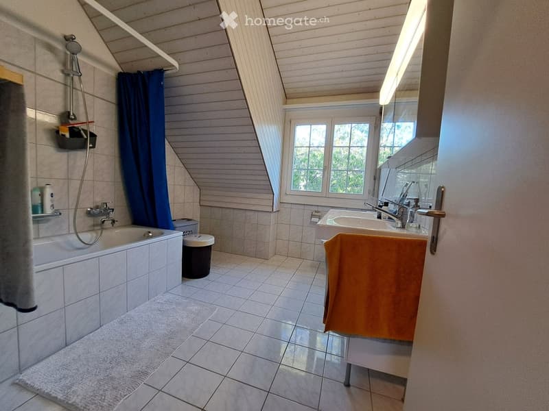Bright bathroom with a bathtub, sink, and mirror. Features a window with a view of greenery, a shower curtain, and a towel on the sink counter.