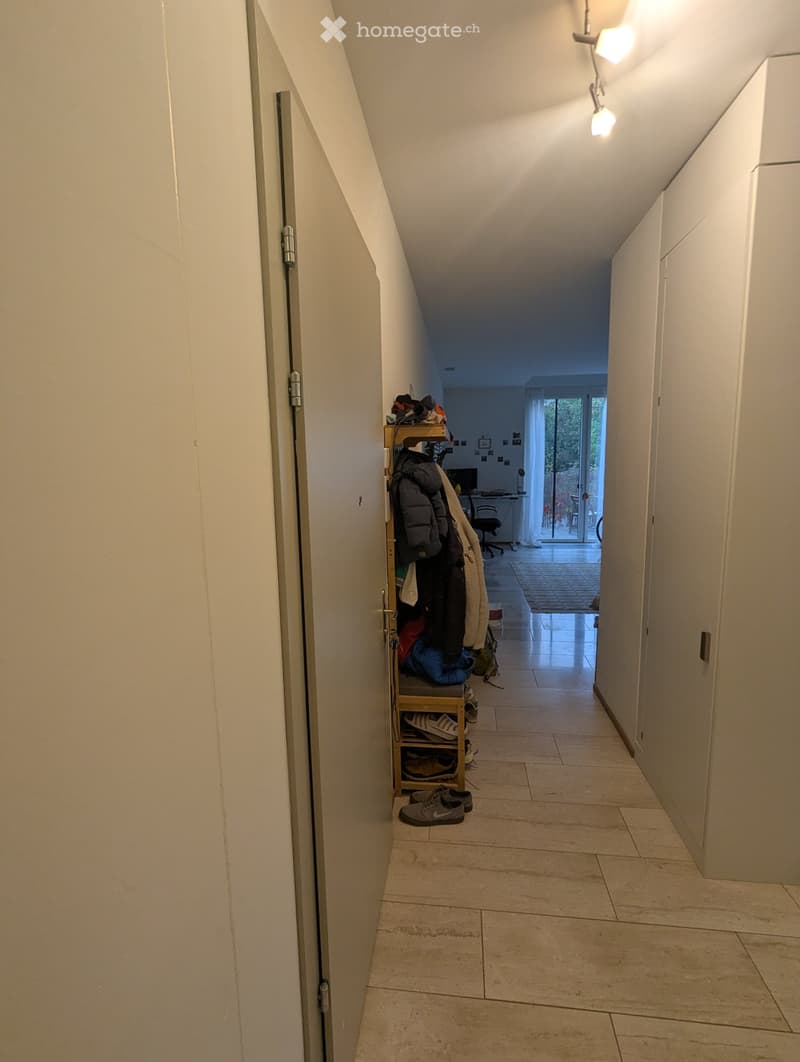 A narrow hallway with tiled flooring, white walls, and built-in storage shelves holding shoes and jackets. The hallway leads to a room with a desk and a sliding glass door to an outdoor area.