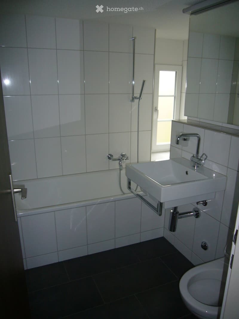 Modern bathroom with white tiled walls, a bathtub, a sink with a chrome faucet, a toilet, and a large mirror above the sink. The room has dark tiled flooring and a window allowing natural light.