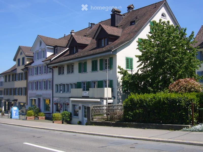 Multi-story residential building with white walls, green shutters, and a brown tiled roof. Features include a small balcony, a fenced garden area, and a storefront with signage on the ground floor.