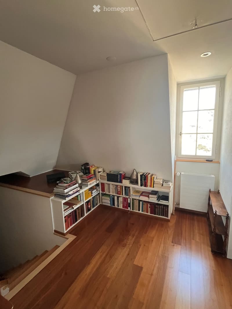 A cozy attic room with wooden floors, a large window, a bookshelf filled with books, and a small table near the window.