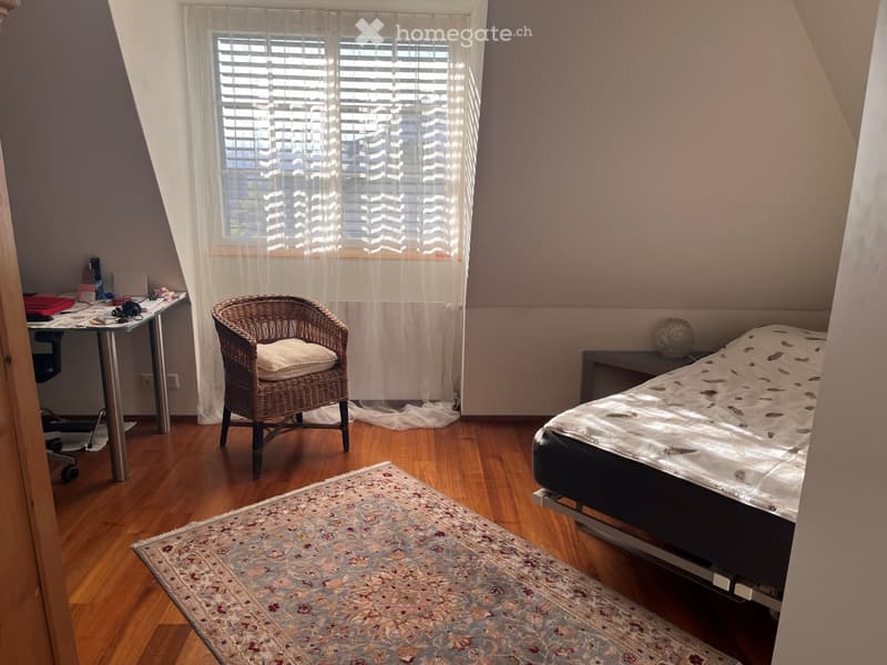 Cozy attic bedroom with wooden floors, a large window with blinds, a bed with patterned bedding, a wicker chair, a desk, and a nightstand.