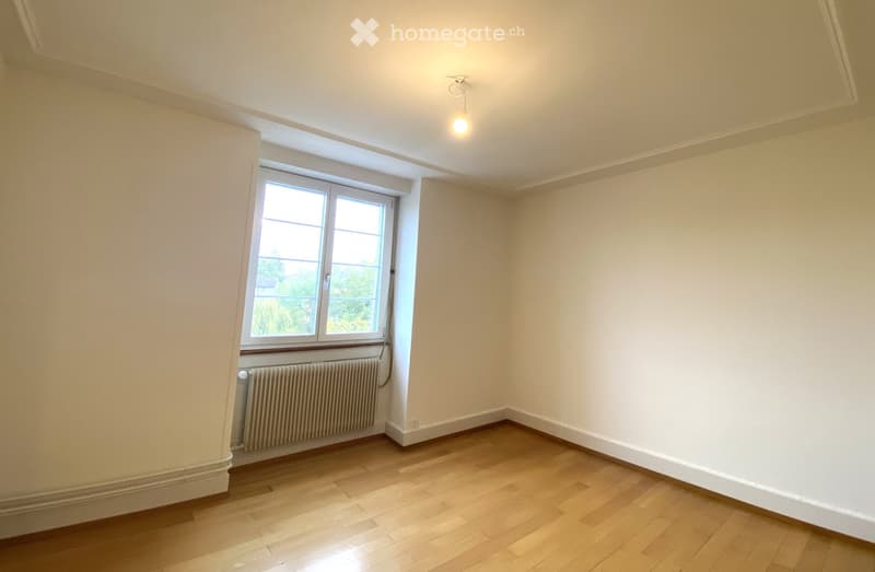 Empty room with wooden flooring, white walls, a single window with a radiator below, and a ceiling light fixture.
