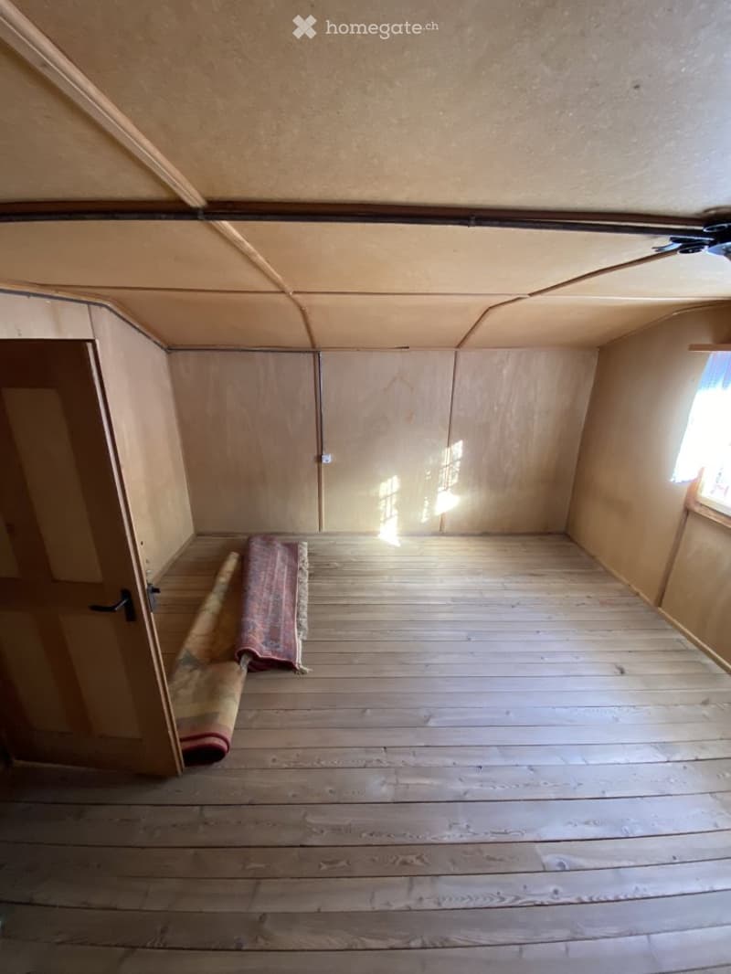 Empty room with wooden flooring, bare walls, and a small window allowing natural light. A rolled-up rug is placed near the door.