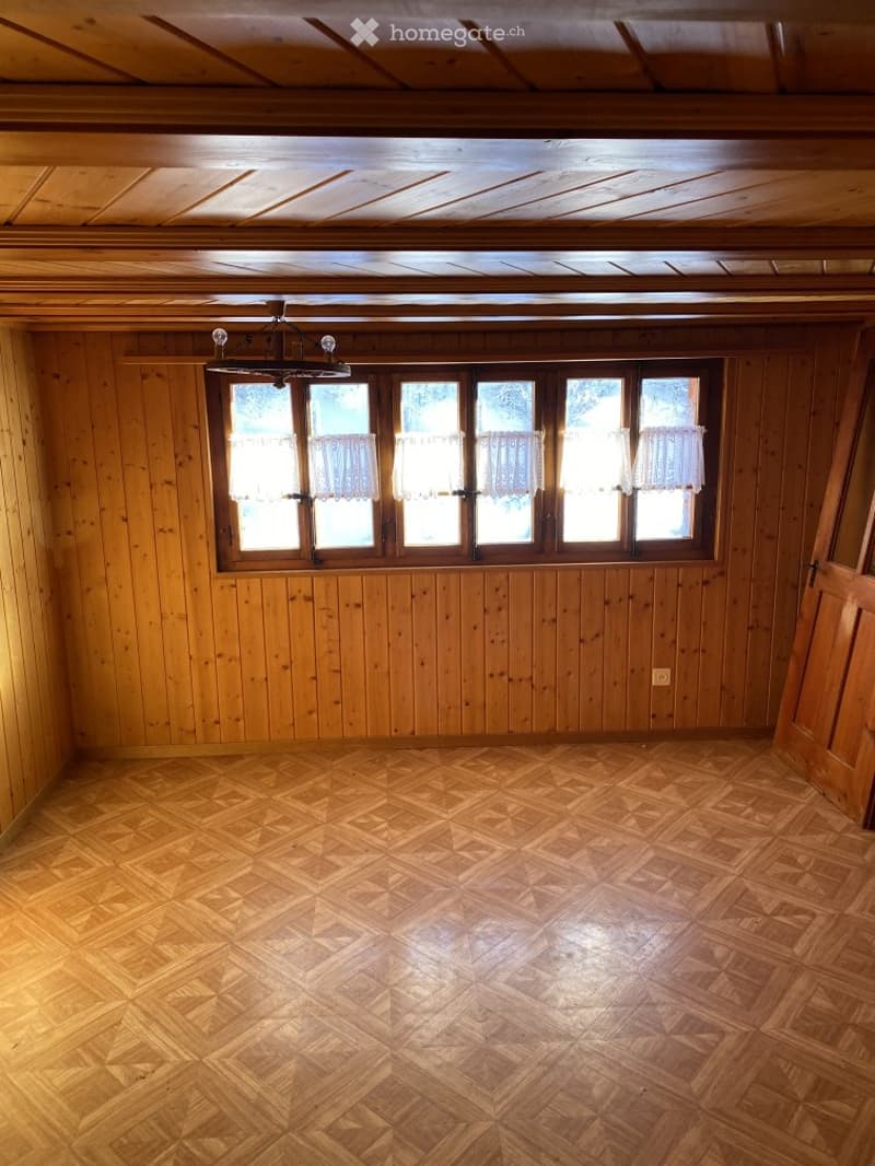 Empty room with wooden walls, wooden floor, large window with curtains, and a chandelier.