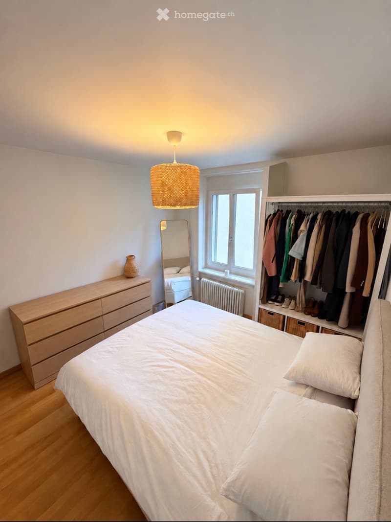 Bright bedroom with a neatly made bed, a wooden wardrobe with hanging clothes, a mirror, a pendant lamp, and a small wooden dresser with a decorative vase on top.