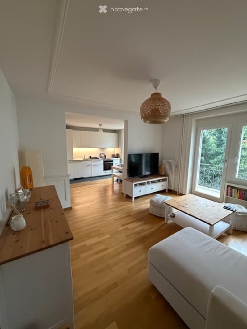 Bright, open-plan living and dining area with wooden flooring, white walls, and large windows offering a view of greenery. Features a modern sofa, coffee table, TV stand, and a small dining area with a table and chairs. The adjacent kitchen has white cabi