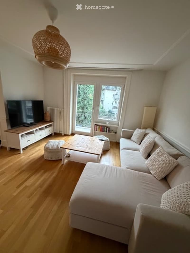 Bright living room with wooden flooring, large window offering a view, a grey sectional sofa, a white coffee table, an ottoman, a TV stand, and a woven hanging light fixture.
