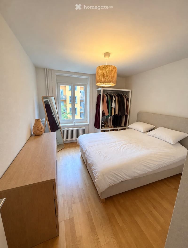 Cozy bedroom with a single bed, white bedding, a wardrobe with hanging clothes, a hanging woven lamp, a mirror, and a decorative vase on a wooden side table.