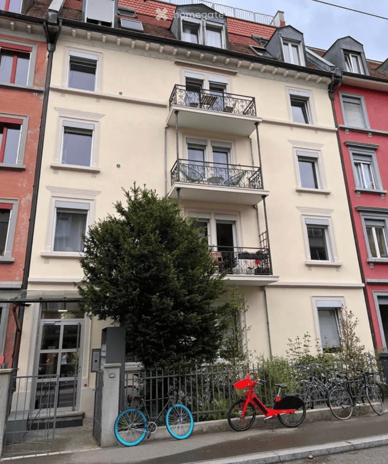Multi-story residential building with balconies, large windows, and a tree in front. Bicycles are parked on the sidewalk.