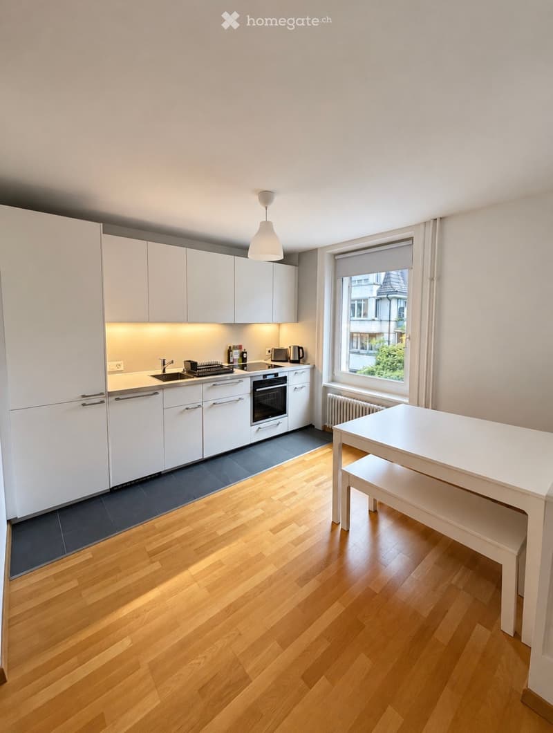 Modern kitchen with white cabinets, wooden flooring, a sink, oven, microwave, and a small dining table with a stool.