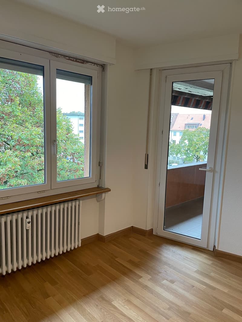Empty room with wooden flooring, large windows, radiator, and sliding glass door leading to a balcony.