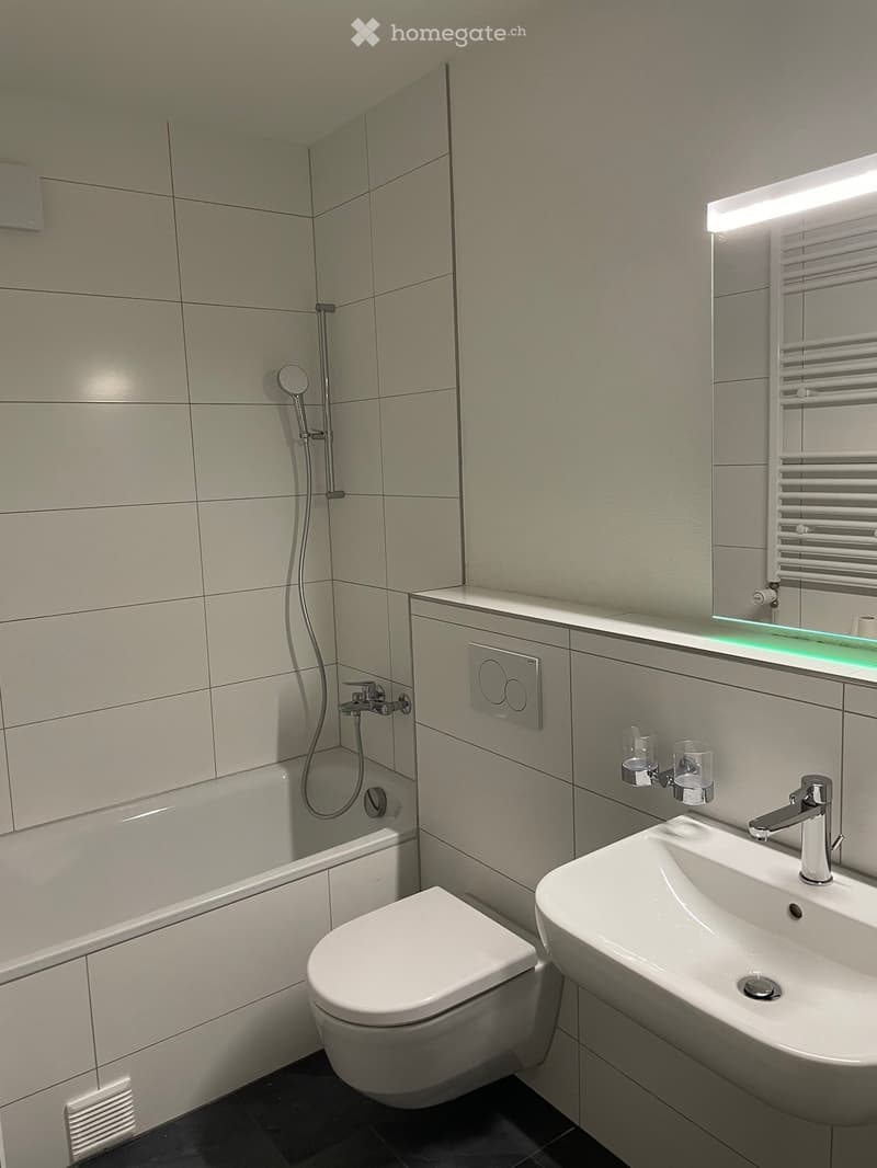 Modern bathroom with a white toilet, sink, and shower. Features a large mirror with built-in lighting, a radiator, and sleek fixtures.