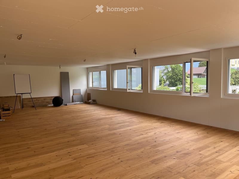 Empty room with wooden flooring, large windows, and minimal furnishings including a whiteboard, a ball, and some stacked items.