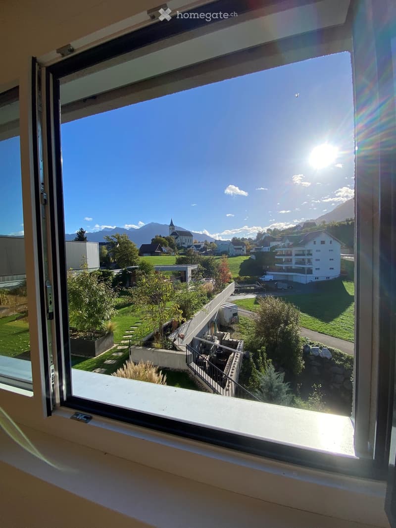 View from a balcony showing a green garden area, a staircase, and a town with houses, trees, and mountains in the background under a clear blue sky.