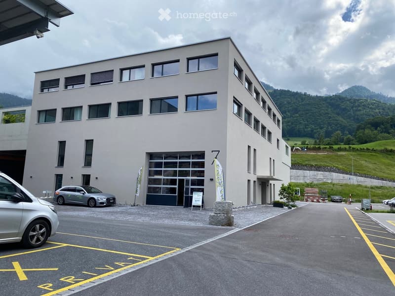 Modern two-story building with large windows and a garage entrance, surrounded by a parking lot with several cars and a scenic mountain backdrop.
