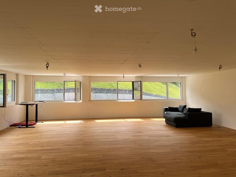 Bright, open living space with wooden flooring, large windows, a black sectional sofa, and a small table with books.