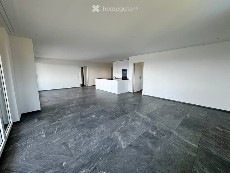 Empty room with grey tiled floor, white walls, and a small white kitchen area visible in the background.