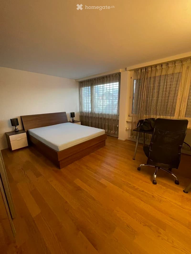 Spacious bedroom with wooden flooring, a large bed with a headboard, a nightstand with a lamp, a chair, and a desk area with a chair and a bag.