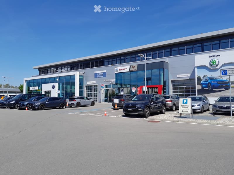 Modern car dealership building with large glass windows and signage for Volkswagen, Seat, and Skoda. Several cars are parked in front, and there is a parking sign visible.