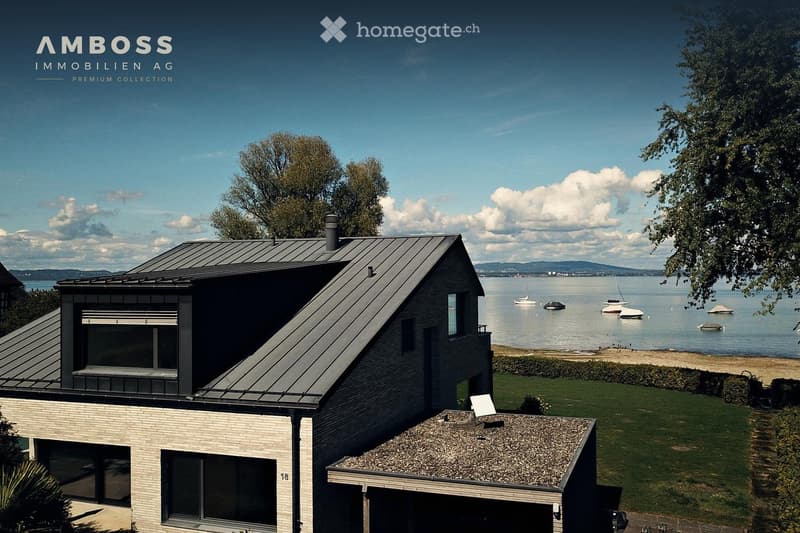 Modern two-story house with a flat roof and large windows, situated near a waterfront with boats visible in the background.