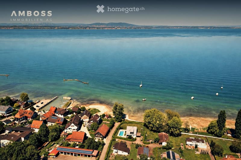 Aerial view of a lakeside residential area with houses, a sandy beach, a swimming pool, and several boats on the lake.