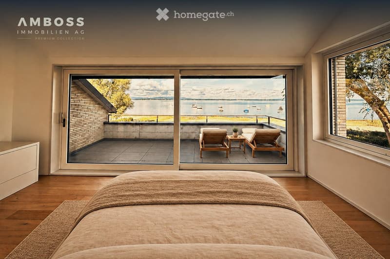 Modern bedroom with wooden flooring, large windows offering a scenic lake view, and a balcony with lounge chairs.
