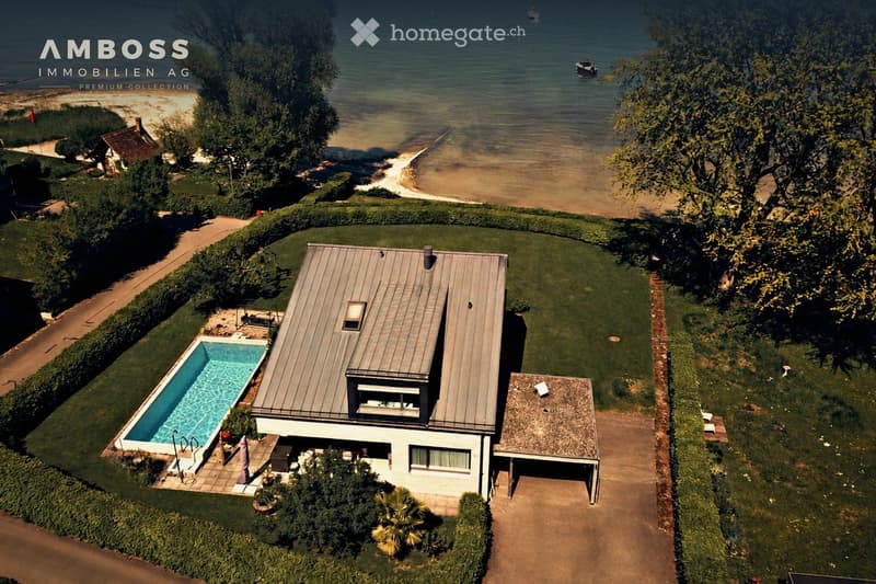 Aerial view of a modern house with a swimming pool, surrounded by a well-maintained garden, near a waterfront with visible boats and a sandy beach.