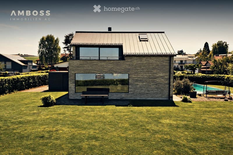 Modern two-story house with a brick facade, large windows, and a flat roof, surrounded by a well-maintained lawn and garden area with a swimming pool.