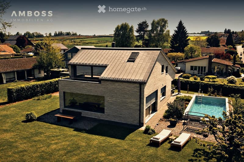 A modern house with a flat roof and large windows, surrounded by a well-maintained garden with a swimming pool, lounge chairs, and a bench.